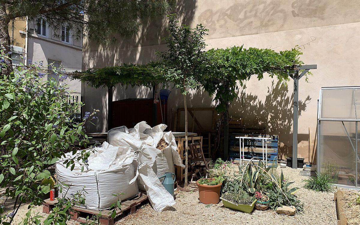 Coin d'un jardin partagé urbain à Lyon, avec pergola, arrosoir, serre et sacs de compost